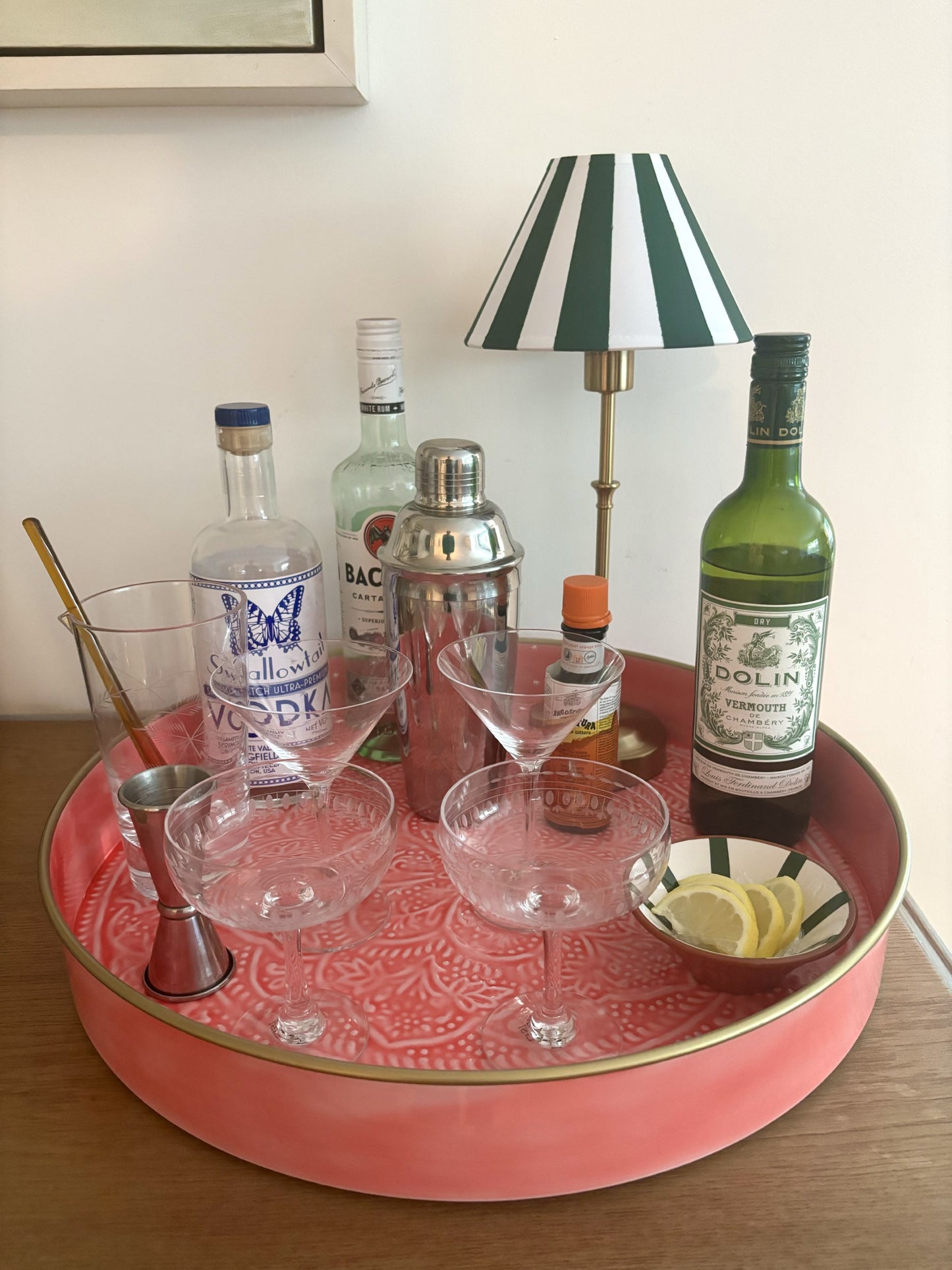 A pink tray with cocktail glassers, a cocktail shaker and spirits.  There is also a cordless green and white striped lamp.