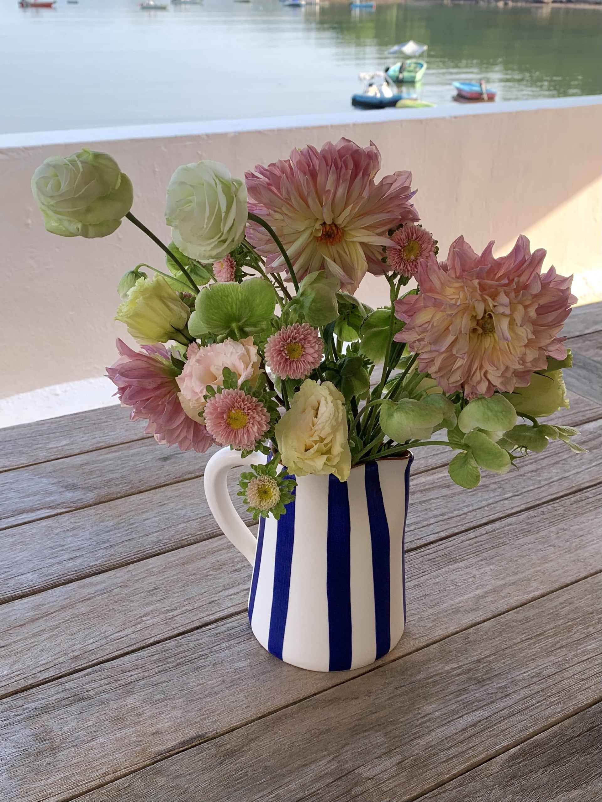 A blue and white hand-painted jug filled with fresh flowers.  The jug is on a wooden table next to the sea.
