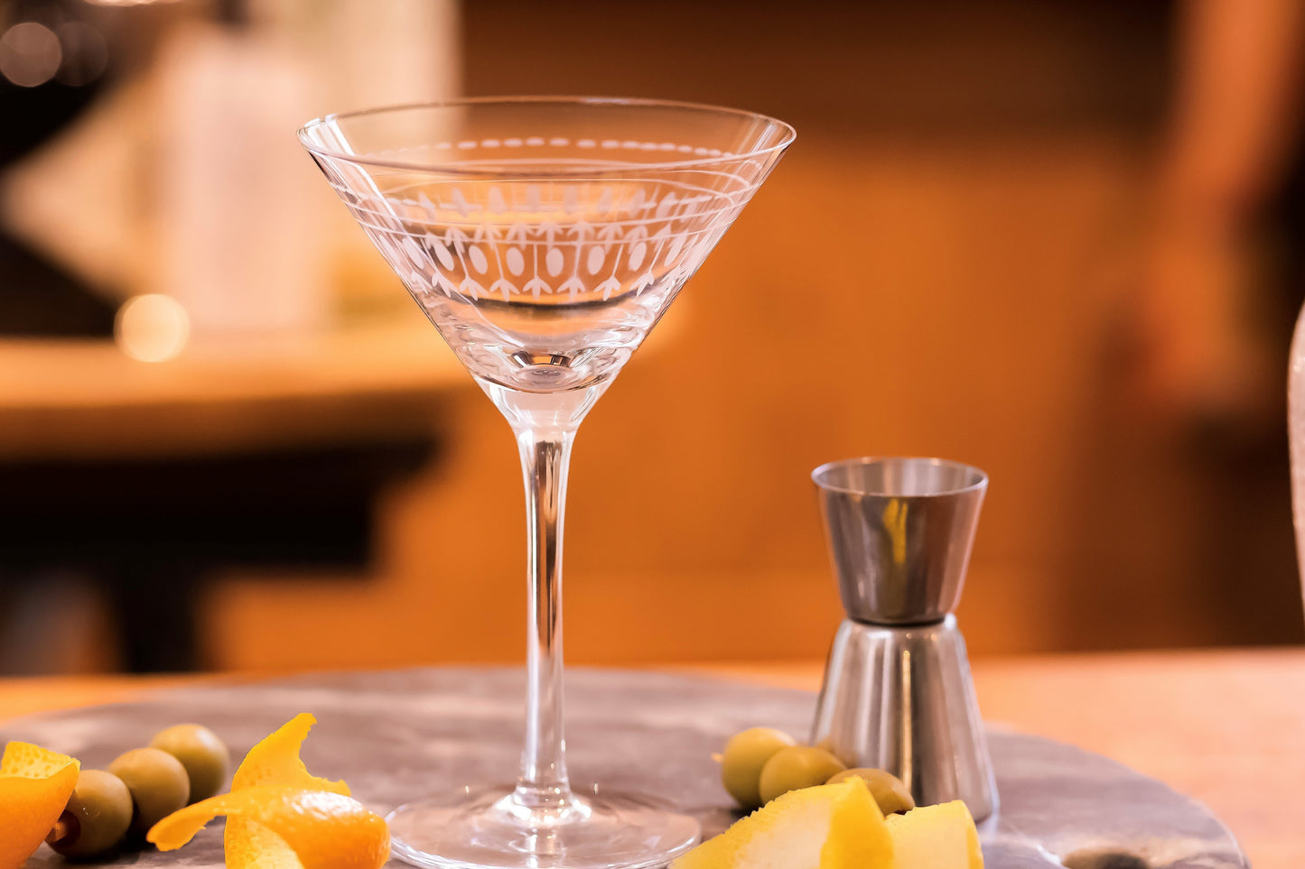 A classic martini glass hand etched with a delicate ovals pattern. There are some olives and orange peel in the background.