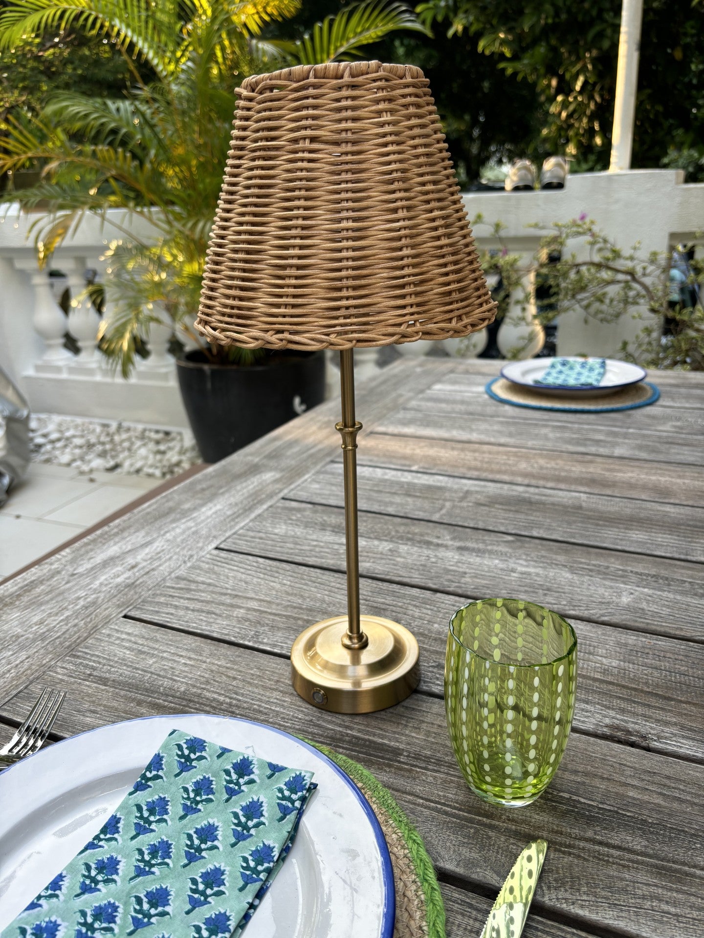 A cordless brass lamp with a rattan shade on an outdoor wooden dining table.