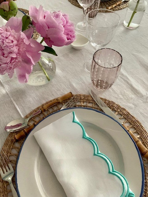 A place setting with a teal linen scallop napkin and a pink glass tumbler with perle design.  There is bamboo cutlery and a vase of pink peonies.
