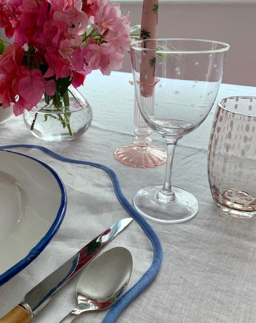 A close up of a place setting with a star etched wine glass and a pink tumbler with perle design.  There is a pink candle in the background and a vase of bougainvillea.