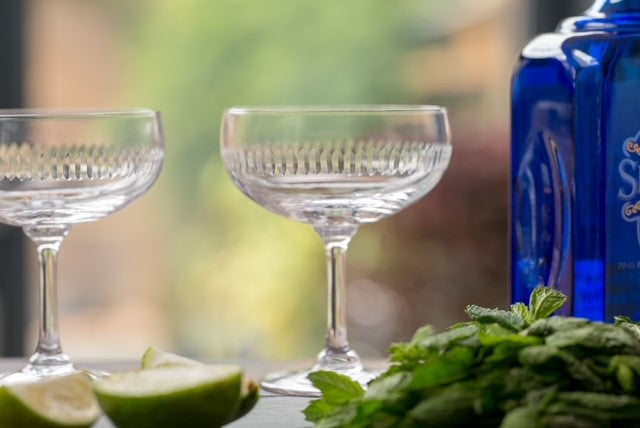 Two cocktail glasses or coupes next to a blue bottle of spirits, a bunch of mint and some lime wedges.