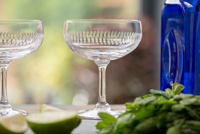 Two cocktail glasses or coupes next to a blue bottle of spirits, a bunch of mint and some lime wedges.