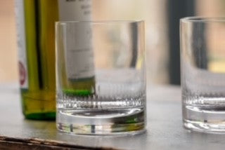 A crystal whiskey glass set next to a bottle. The whiskey glass has an engraved spears pattern around the base rim.