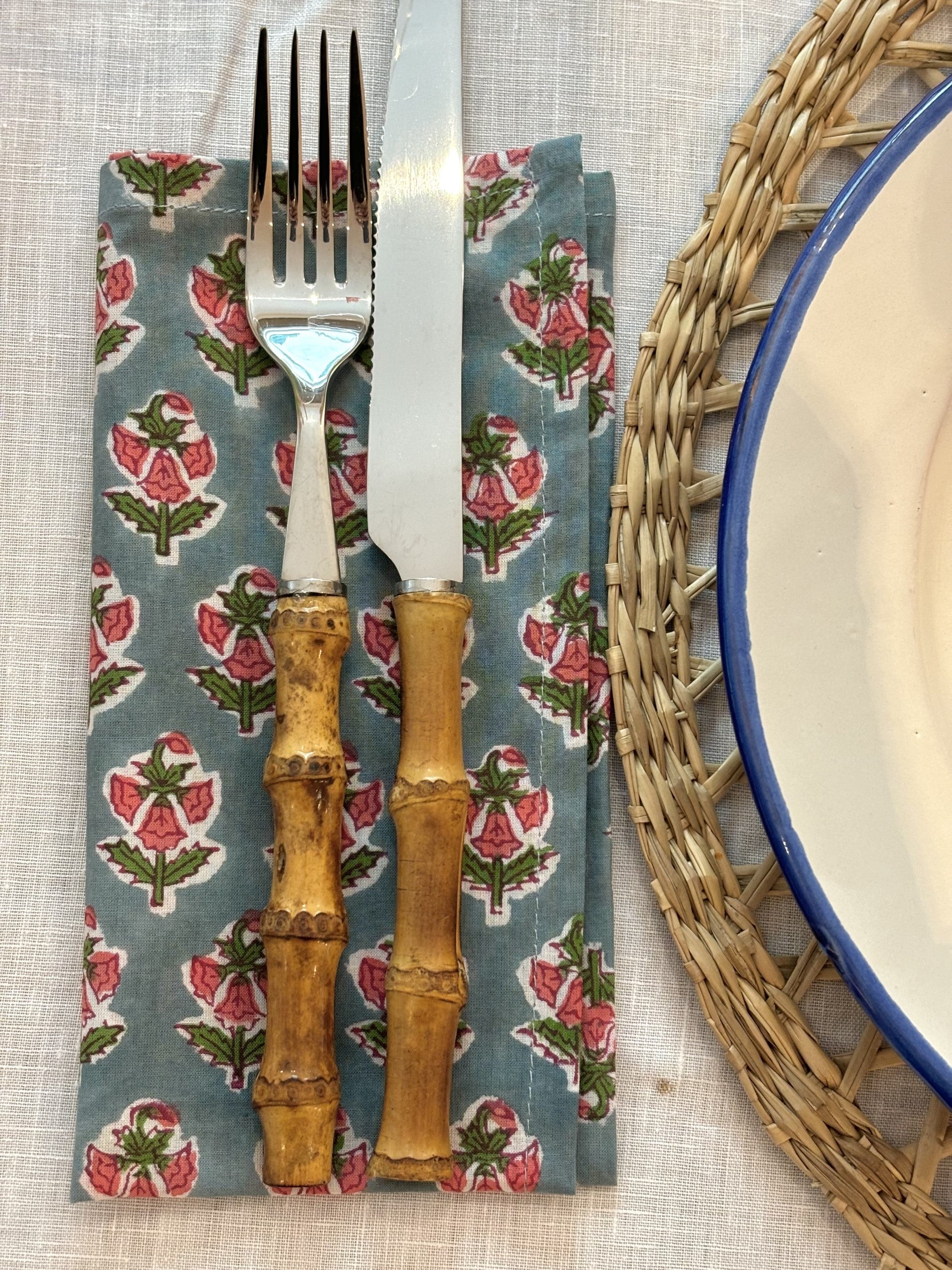 A blue and pink floral block printed napkin with bamboo cutlery on top next to a placemat.