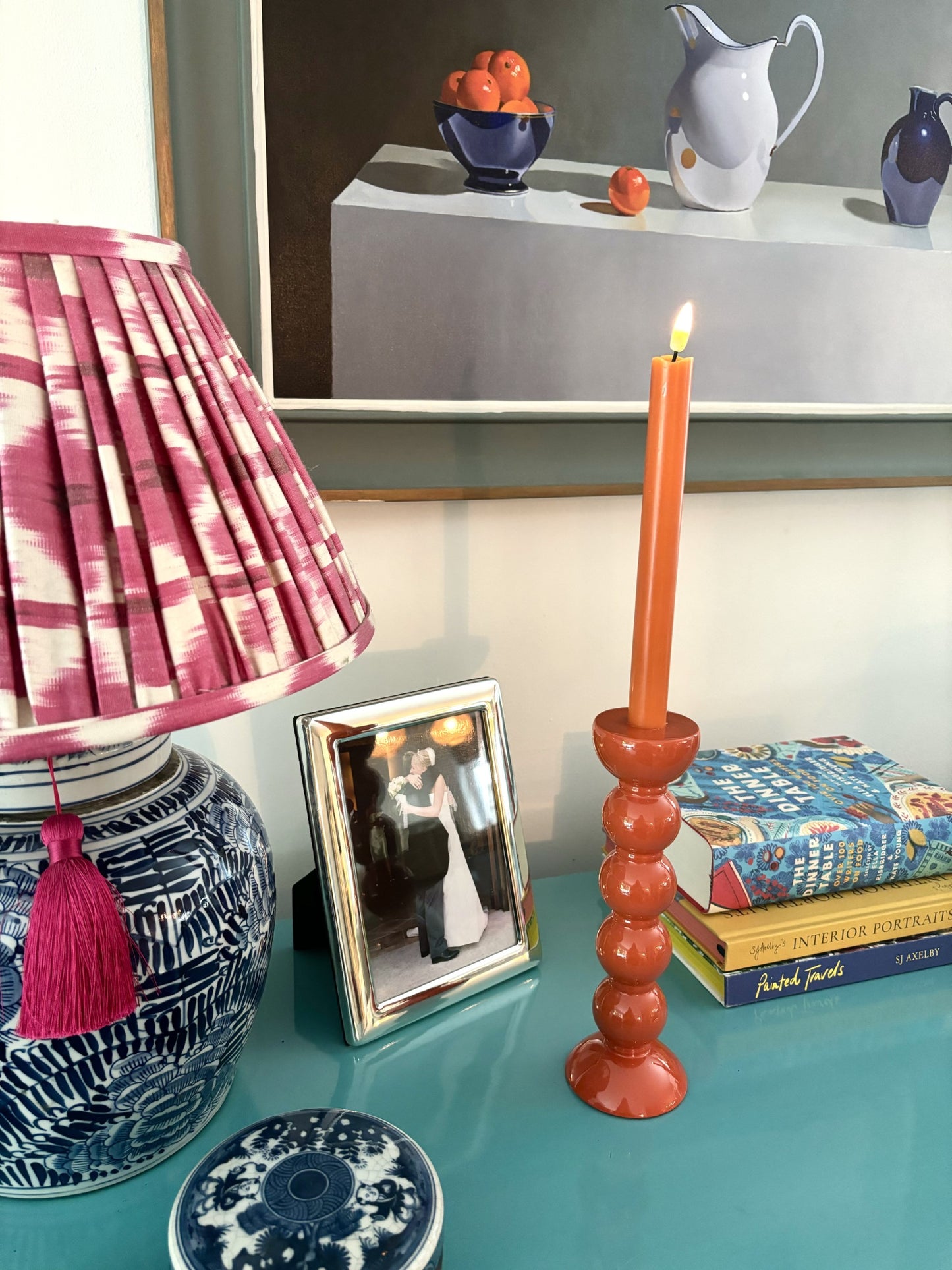 An orange bobbin candlestick on a sideboard with a ginger jar lamp, photo and books.  There is a still life painting of a bowl of oranges in the background.