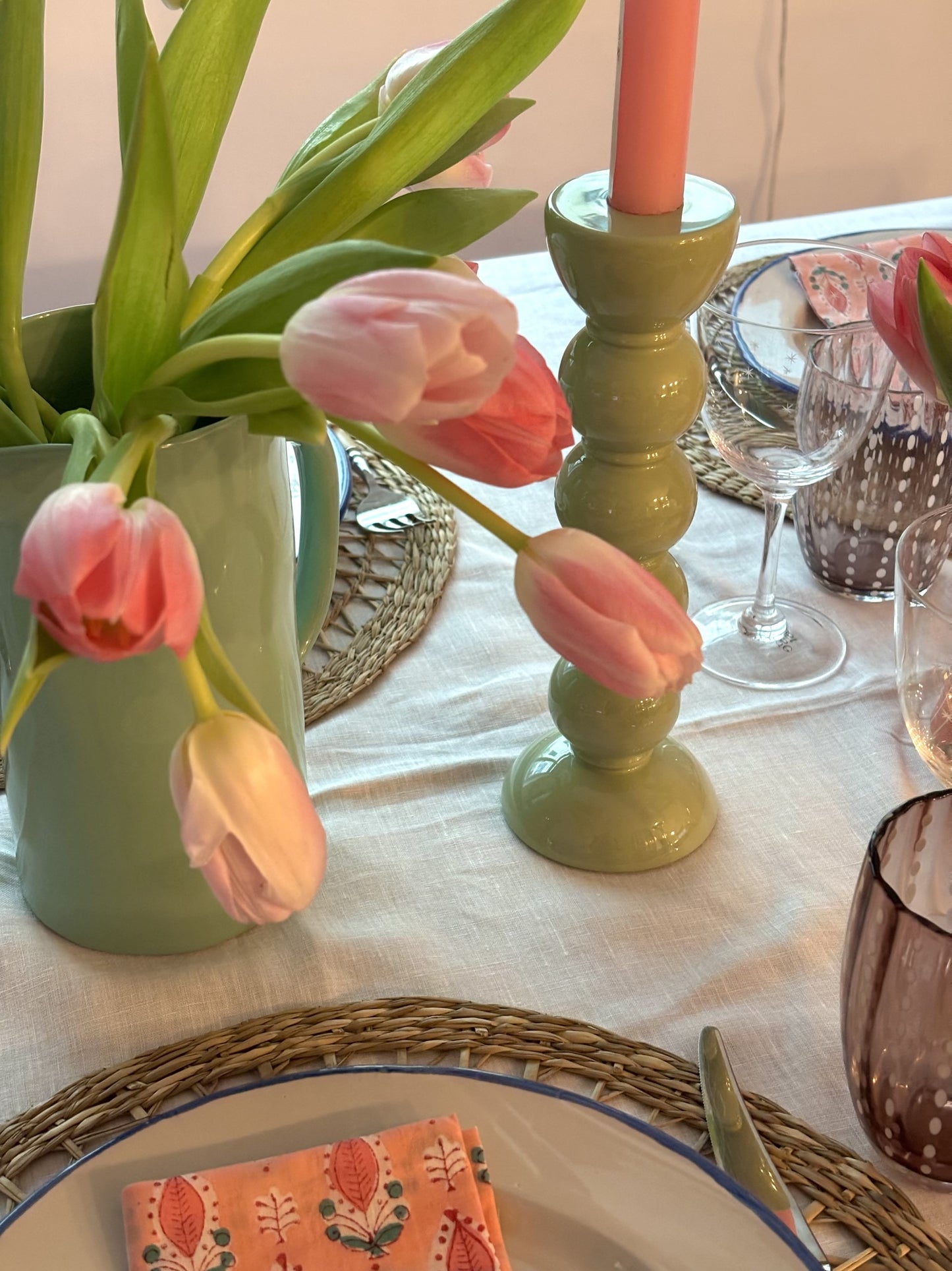 A close up of a sage green bobbin candlestick with a rose candle next to a jug of pink tulips.
