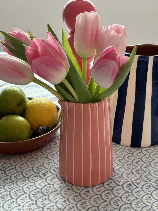 A small pink and white striped milk jug filled with pink tulips next to a bowl of fruit.