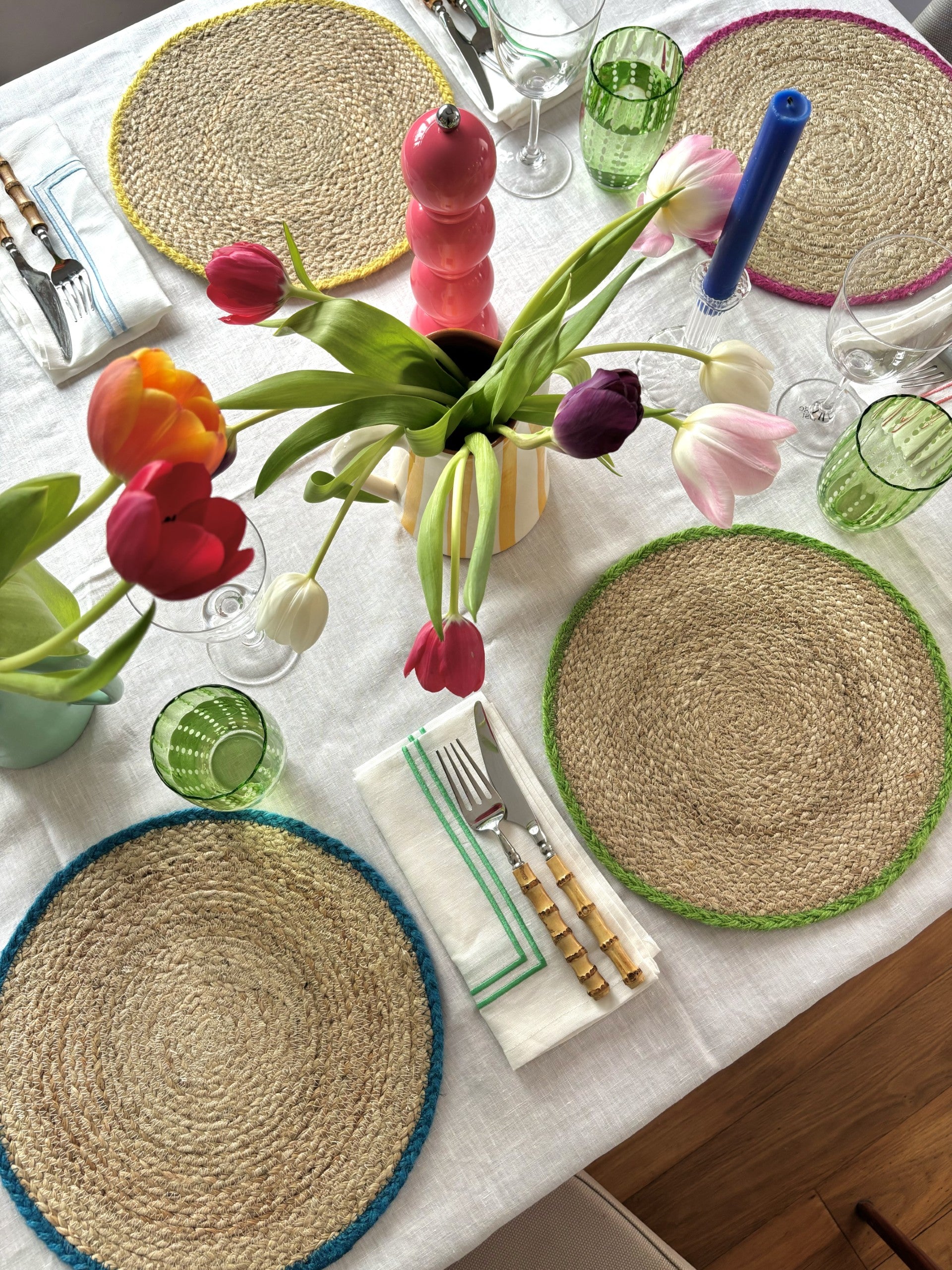 A tablescape with jute placemats with a colourful border, tulips in jugs and green glass tumblers. 