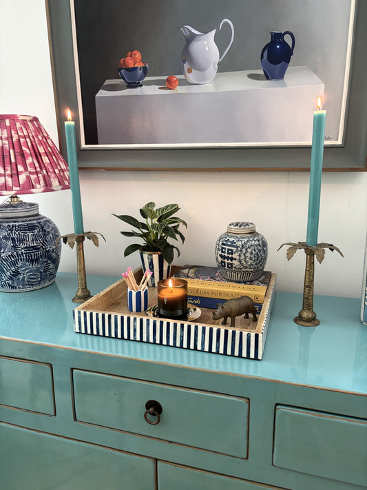 A styled blue and white bone inlay tray on a blue sideboard under a painting of a some jugs and oranges.