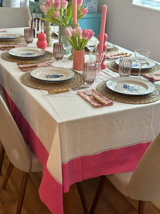 A laid table with a white linen tablecloth with a bright pink border.  There are jugs of fresh tulips on the table and pink candles.