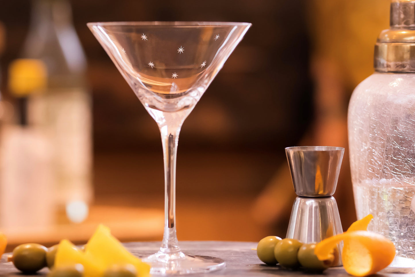 A classic martini glass with an engraved stars pattern.  Next to a cocktail shaker and some olives.