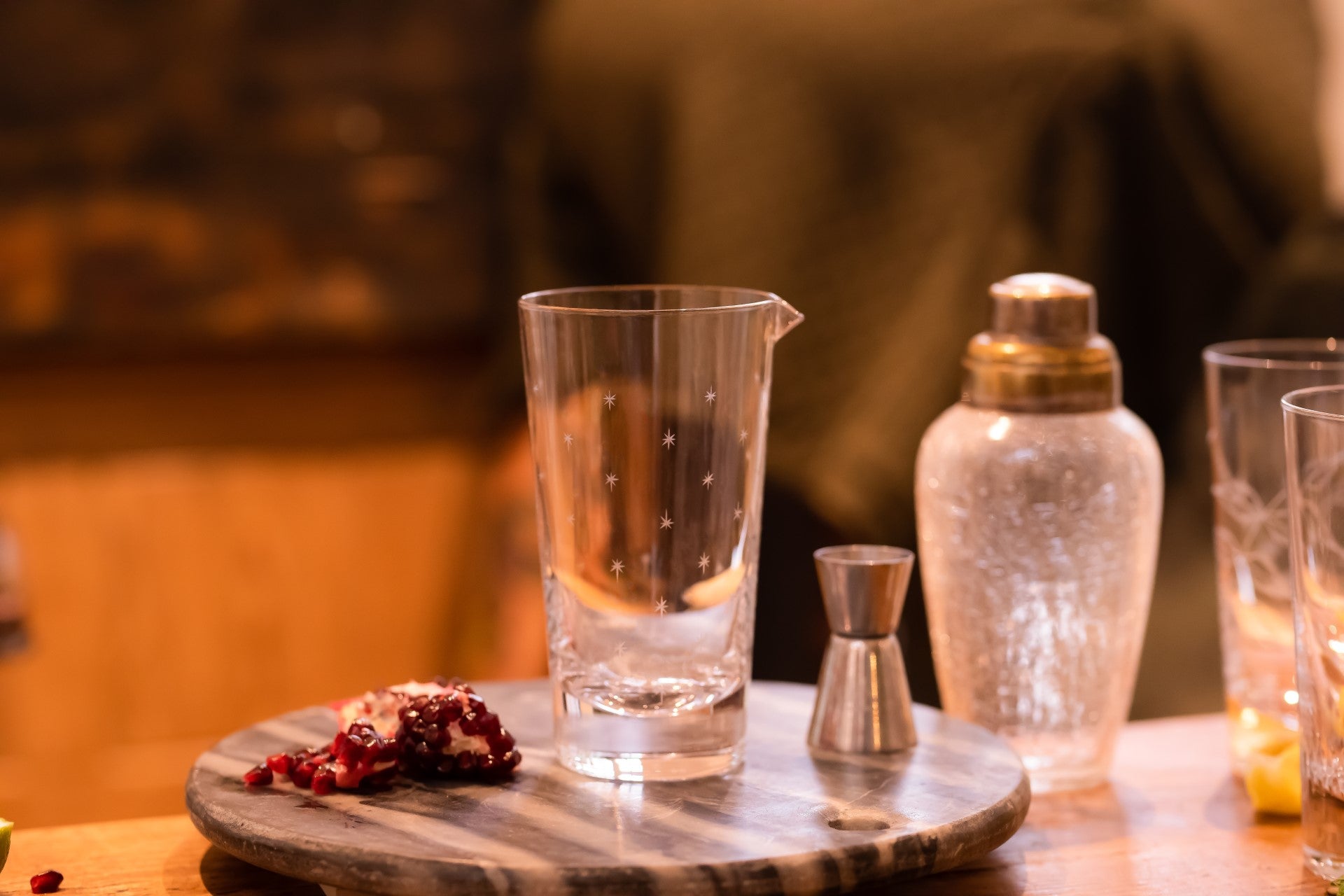 A crystal mixing glass with a stars design sitting on a board next to a cocktail shaker and glasses.