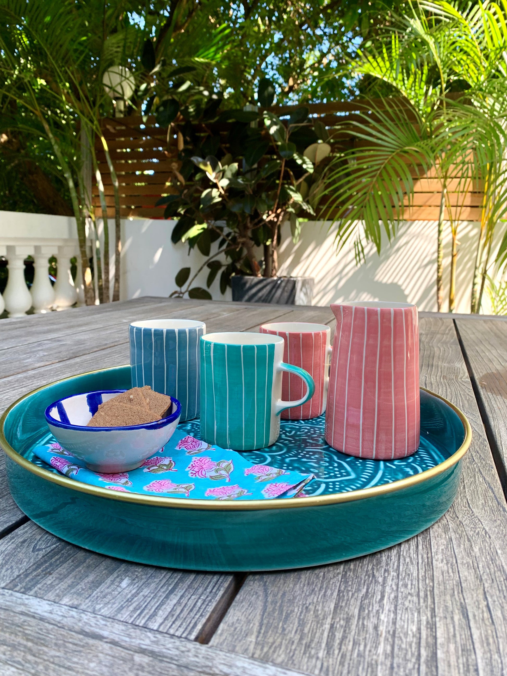 A teal tray on a table in the garden with some coffee mugs, a milk jug and a block printed flowery napkin.