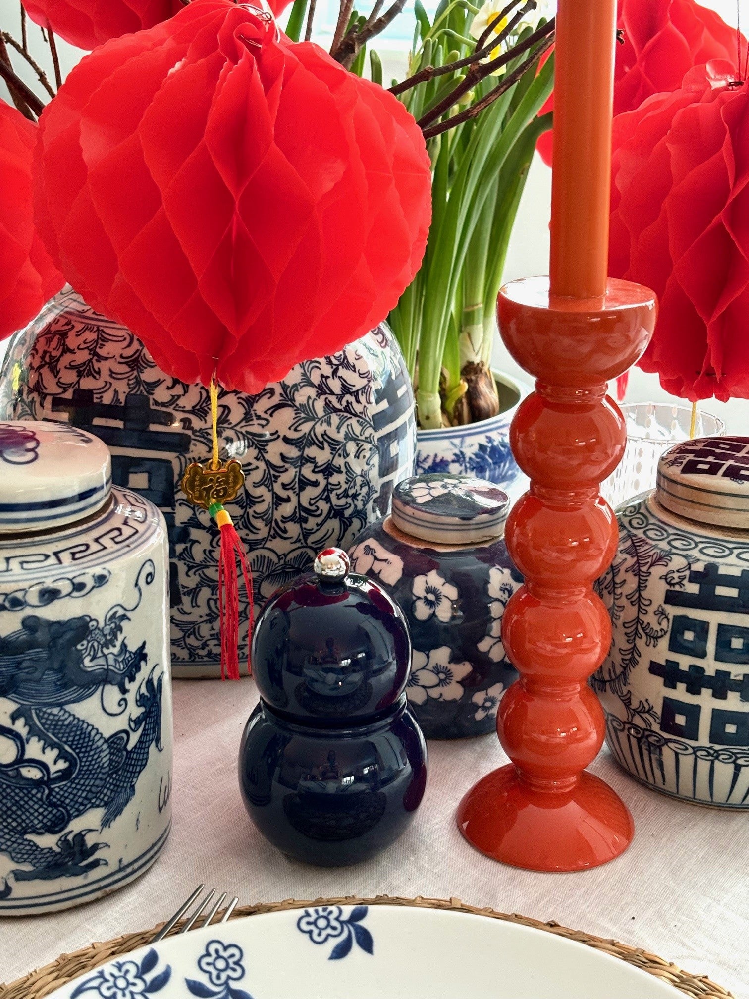 An orange bobbin candlestick on a table surrounded by blue and white ginger jars and red lanterns.