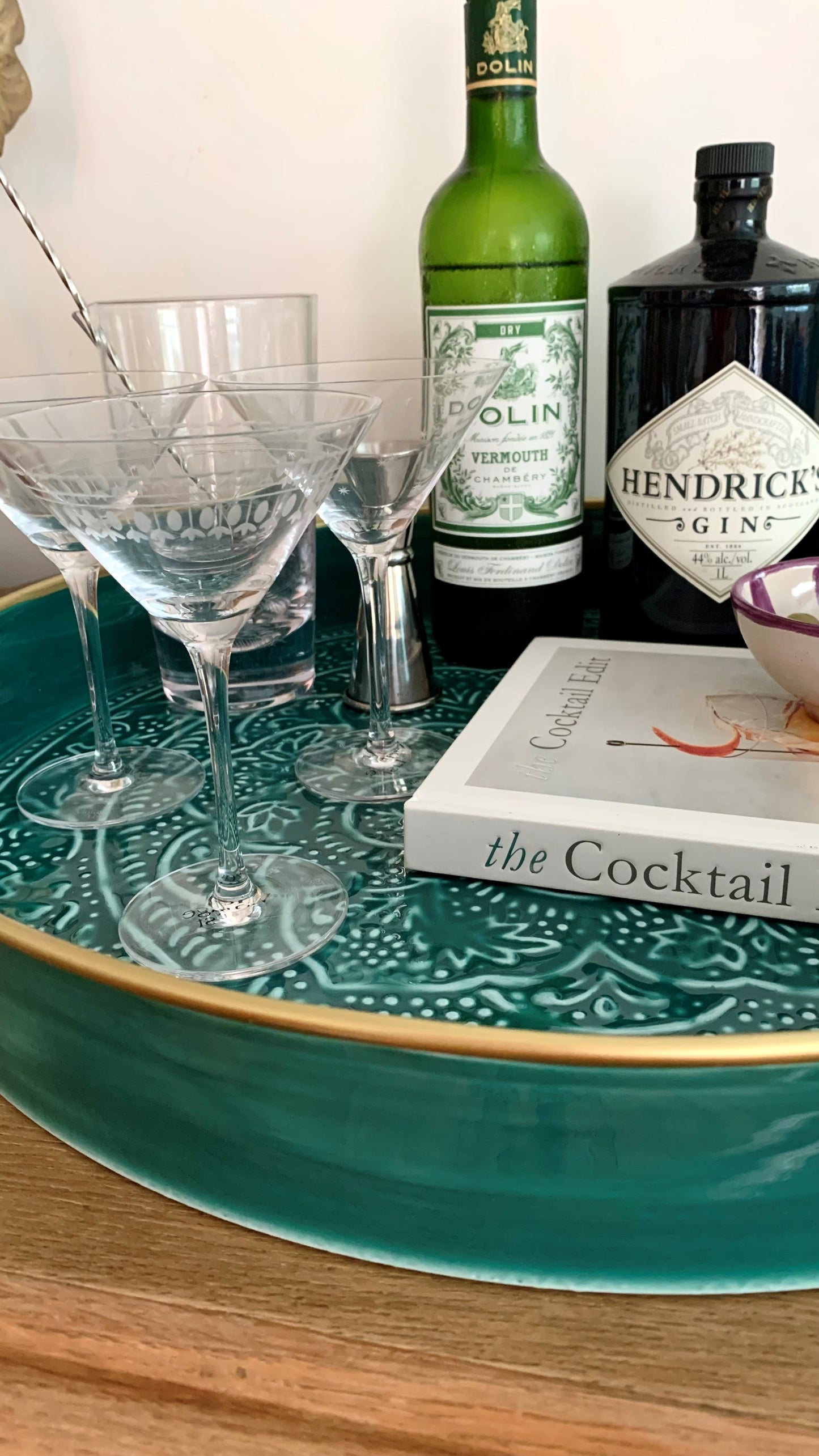 A teal drinks tray with a cocktail book, spirits, a cocktail mixing glass and 3 hand-etched crystal martini glasses