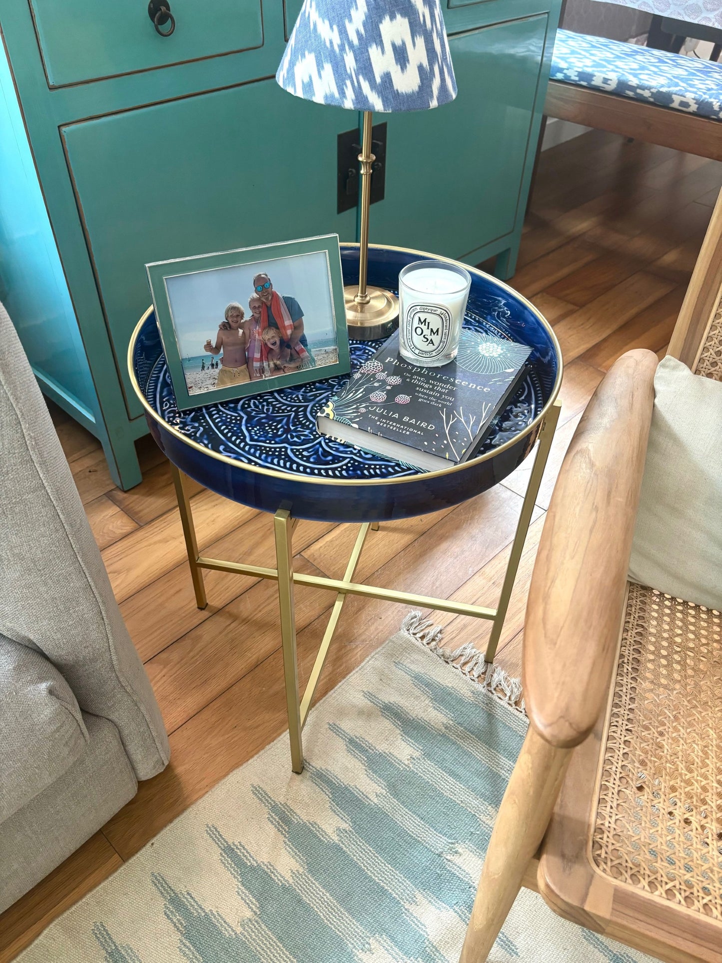 A large blue tray on a brass stand used as a side table next to a sofa.  There is a lamp, photo, book and candle on the tray.