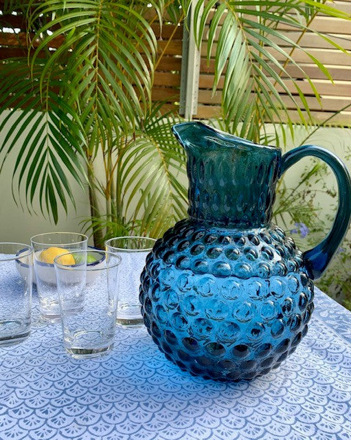 A large blue hobnail jug outside on a table next to 4 glass tumblers and a bowl of lemons.  There is a palm tree in the background.