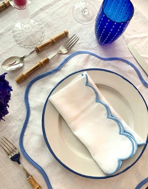 A blue and white place setting.  White linen placemats with a blue scalloped border paired with white linen napkins with a blue scalloped edge.  There is bamboo cutlery and a blue glass perle tumbler.