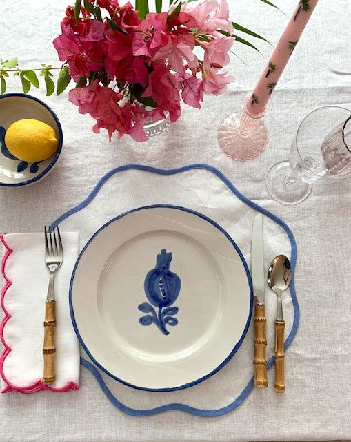 A place setting featuring a blue scalloped placemat and a pink scalloped napkin. There is a vase of pink bougainvillea on the table next to a pink candle and a bowl of lemons.