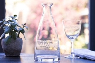 A crystal carafe with a delicate engraved ovals pattern near the base.  The carafe is set on a table next to a crystal wine glass and a potted plant.