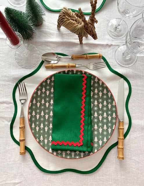 A Christmas place setting with a white linen scallop placemat with a green border and green linen napkins with a red ric-rac edging.  There is bamboo cutlery, 2 small christmas trees and a wicker reindeer on the table.