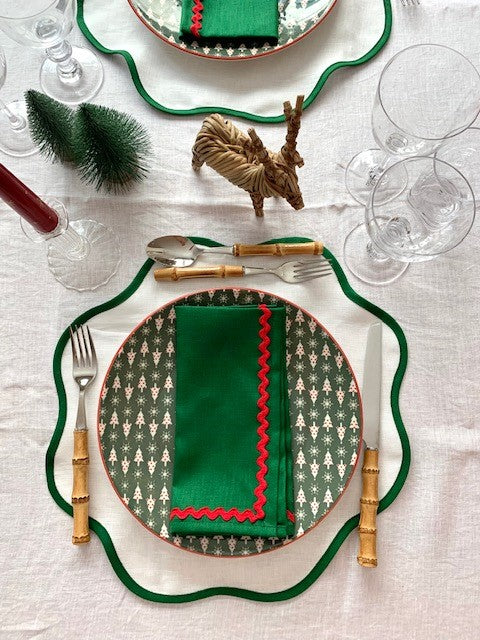 A birds-eye view of a Christmas table setting.  White linen placemats with a green border are paired with green linen napkins with a red ric-rac edging.  There is bamboo cutlery on the table and two small Christmas trees and a wicker reindeer.
