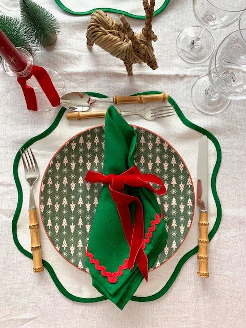 A Christams place setting featuring white linen scallop placemats with a green border and green linen napkins with red ric-rac edging. Ther is also bamboo cutlery, wine glasses, a red star candle and a wicker reindeer.