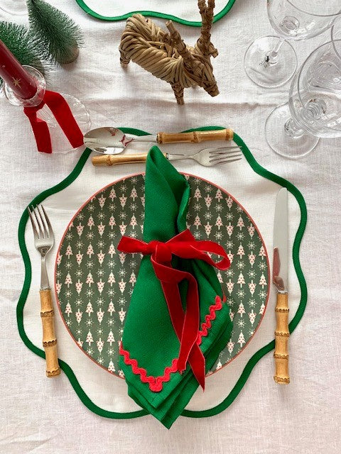A white, green and red Christmas place setting with scallop placemats and green linen napkins.  There is a wicker reindeer and a red candle along with some glassware and bamboo cutlery.