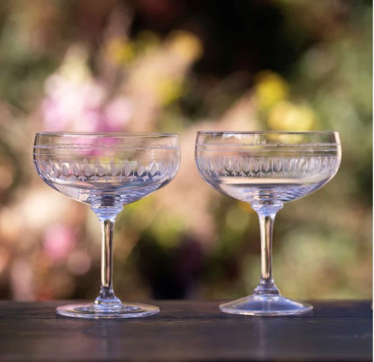 Two crystal cocktail glasses or coupes set on a table outside.  Each glass has been engraved with a delicate ovals pattern.