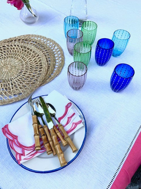 An outdoor dining table with some assorted glass perle tumblers on it.  There is also some white linen napkins with pink scallop edges, bamboo handled cutlery and wicker placemats.