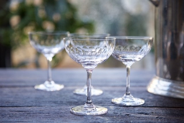 Four vintage inspired crystal champagne glasses next to a silver wine bucket.  Each glass has been engraved with a band of connecting ovals.