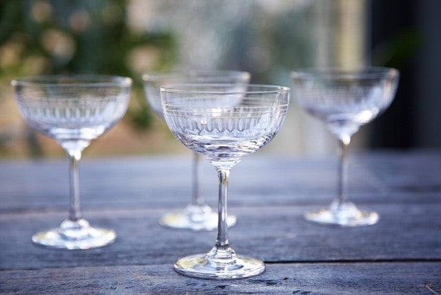 Four vintage inspired crystal champagne glasses sitting on a table.  Each champagne coupe has an engraved ovals pattern.