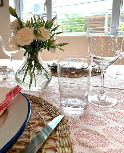 A table setting featuring a crystal glass tumbler and crystal wine glass.  Both glasses feature an engraved star pattern. There is a crystal vase filled with white flowers also on the table.
