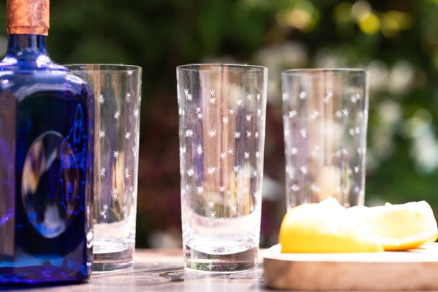 Three crystal highball glasses set on a table outside next to a spirits bottle and some lemon wedges.  The highball glasses have been engraved with stars.