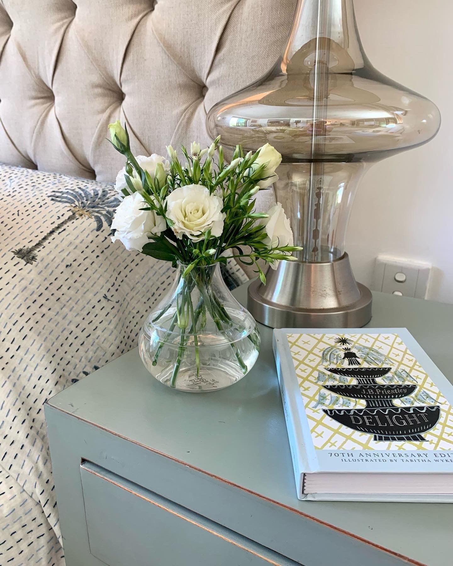 A crystal vase filled with flowers sitting on a bedside table next to a book and a lamp.