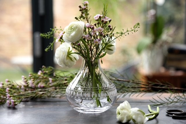 A tear drop crystal vase with an engraved ovals pattern.  The vase is filled with flowers and there are some other flowers lying on the table next to it.