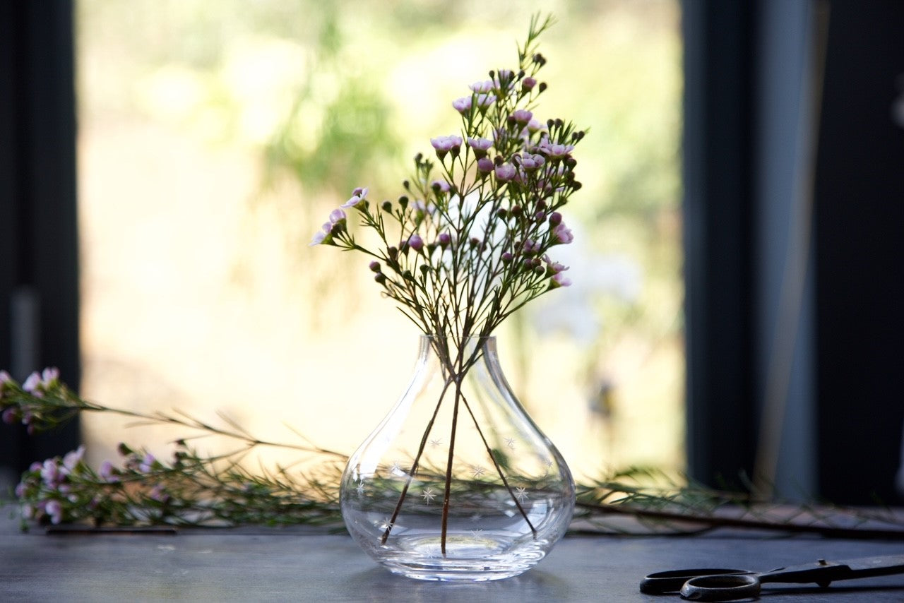 A tear-shaped crystal vase from the vintage list with a hand-etched star pattern. The vase is on a table and is filled with a few flower stems.