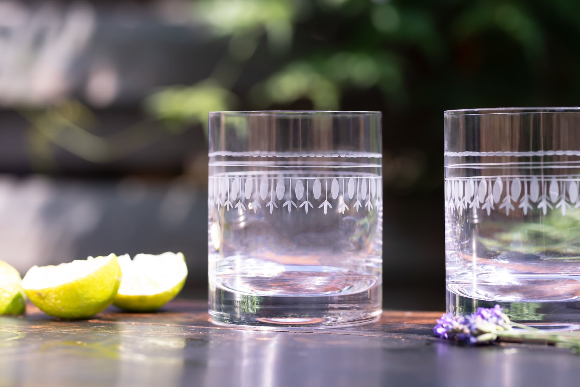 A pair of crystal whiskey glasses outside on a table next to some lemon wedges.  Each whiskey glass has been hand-etched with an oval pattern 