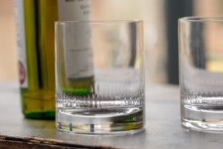 A crystal whiskey glass set next to a bottle.  The whiskey glass has an engraved spears pattern around the base rim.