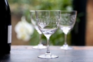 A crystal wine glass with an engraved band of connecting ovals set on a table next to a wine bottle.  There are two other wine glasses in the background.
