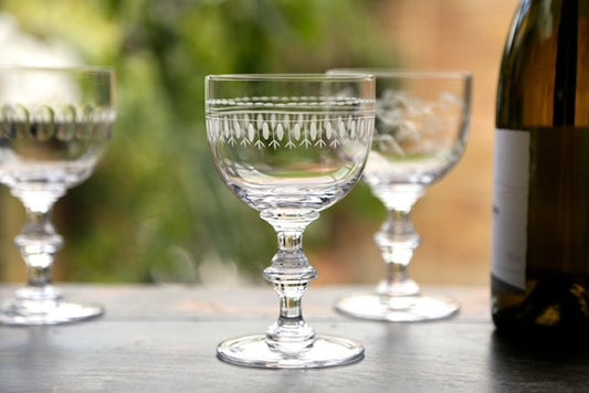 A crystal wine goblet with an engraved ovals design next to a wine bottle.  There are two other wine goblets in the background.