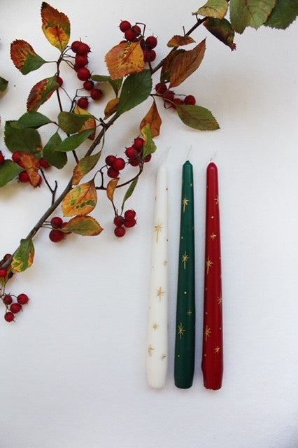 Three Christmas candles lying next to some red berries. One ivory candle hand-painted with gold stars, one green candle hand-painted with gold stars and one red candle hand-painted with gold stars.
