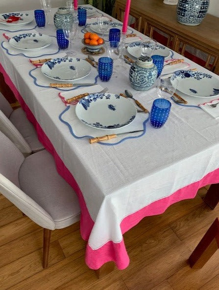 A table setting featuring a white linen tablecloth with a bright pink border.  Placemats, plates and glasses are all blue and white with small ginger jars in the middle of the table.