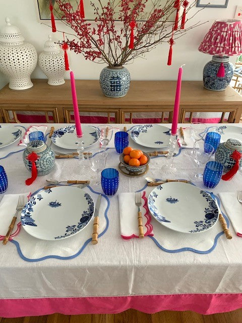 A Chinese New Year table setting featuring a white linen tablecloth with a bright pink border.  The plates, glasses and placemats are all blue and white with double happiness ginger jars in the centre along with a bowl of mini oranges.