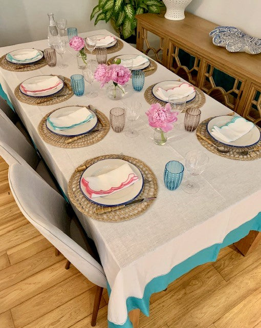 A table setting featuring a white linen tablecloth with a teal border.  The table linen is a mix of teal and pink and there are pink and blue perle glass tumblers.
