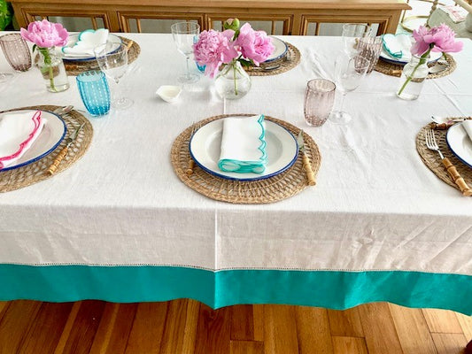 A table setting featuring a white linen tablecloth with a teal border.  Teal and pink scalloped napkins are on the place setting with pink peonies in vases.