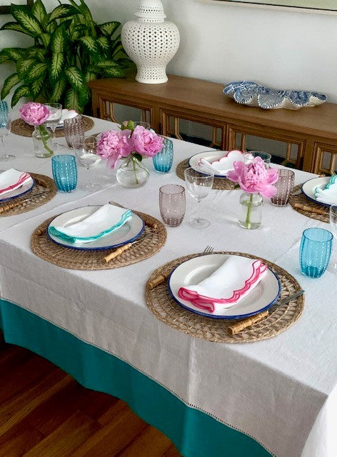 A laid table with white, blue and pink table linen.  The napkins are white with blue or pink scallop edges and the tablecloth is white with a blue border. There is pink and blue perle glassware and small vases of pink peonies.